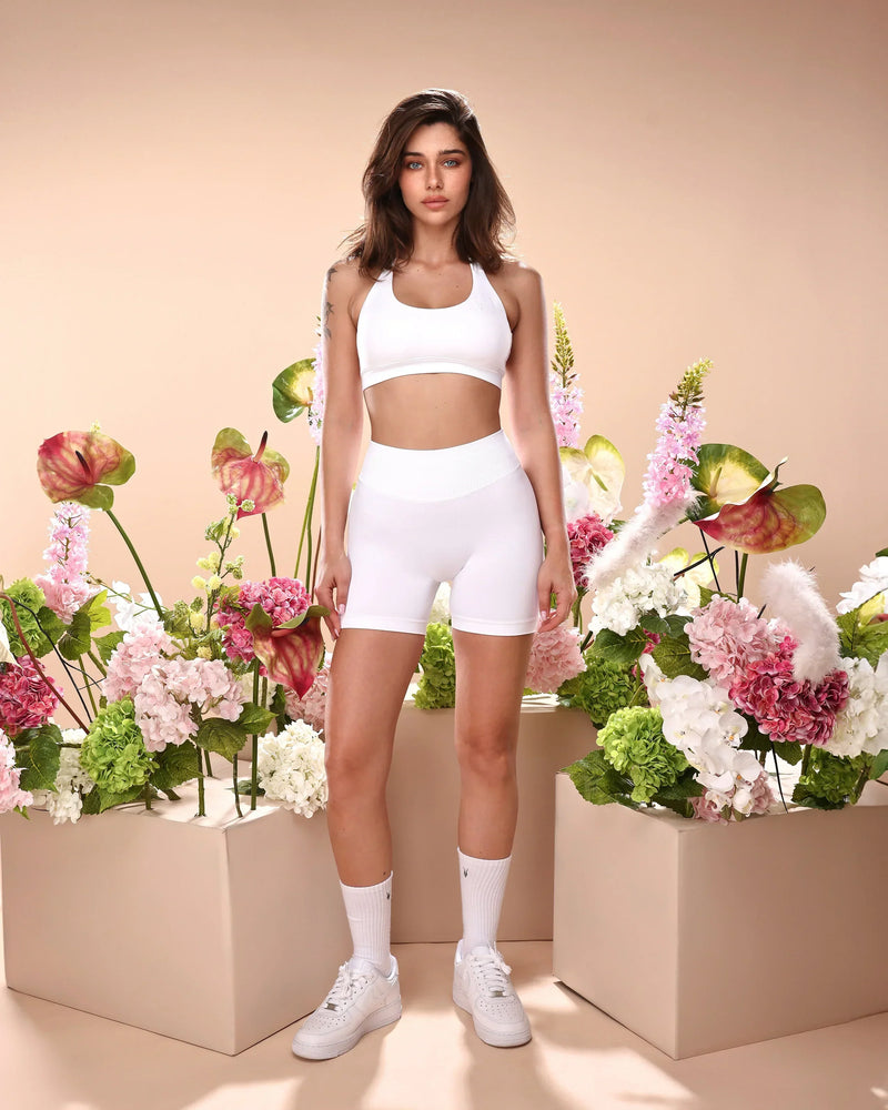 סט בגדי ספורט לנשים בצבע לבן – טופ AURA SNOW ללא תפרים ומכנסיים קצרים תואמים של Inner Dragon. דוגמנית עומדת בסטודיו, מראה את הסט במלואו. מושלם לכושר, יוגה, ריצה ופילאטיס – דגם מומלץ לנוחות, תמיכה ועיצוב מחמיא.