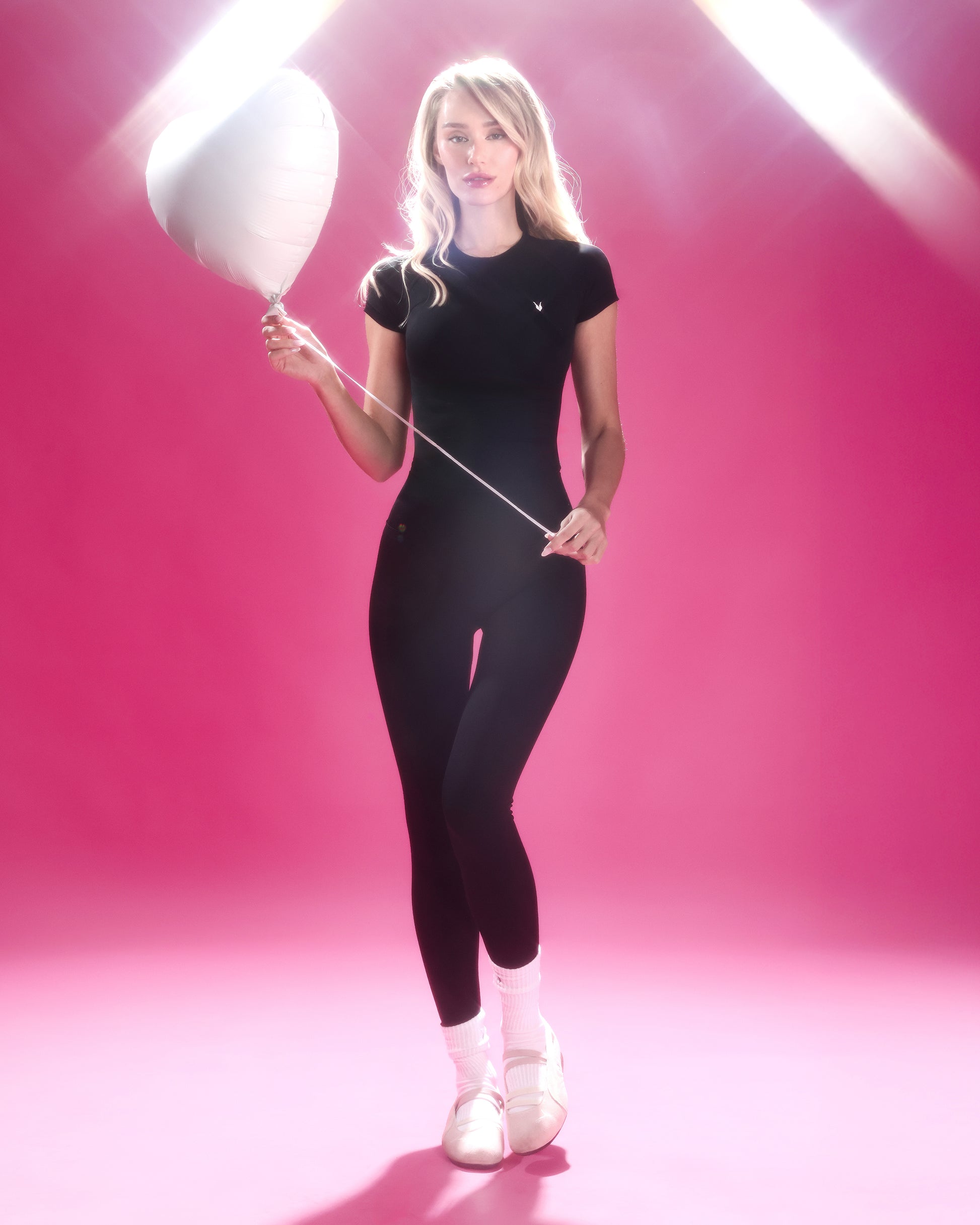 דוגמנית לובשת חולצת ספורט שחורה מחטבת FLORA של Inner Dragon עם טייץ שחור, עומדת בתנועה על רקע ורוד ומחזיקה בלון לבן, מראה נשי ונקי