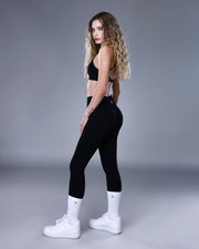 גופיית ספורט NOVA BLACK של Inner Dragon – תצוגת גוף מלאה עם טופ שחור בגב מוצלב (X-Back) ובד אלסטי מחטב. צילום באורך מלא המדגיש את הגזרה הנשית, השילוב עם טייץ שחור מחמיא ונעלי ספורט לבנות.